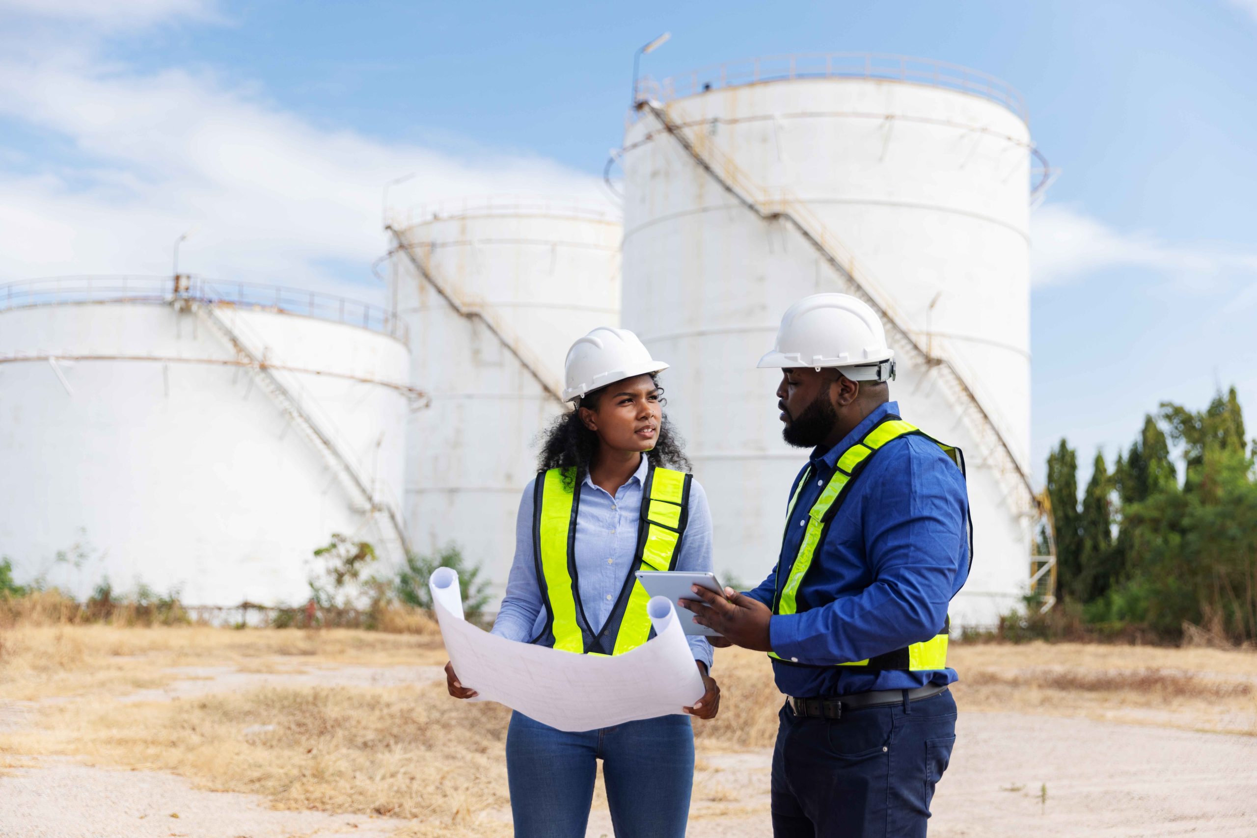 En Grupo Acura, somos tu socio estratégico para implementar soluciones hídricas sostenibles y eficientes a la medida de tu industria. Dos ingenieros o profesionales revisando planos frente a grandes tanques de almacenamiento industrial de Grupo Acura, simbolizando una alianza estratégica.