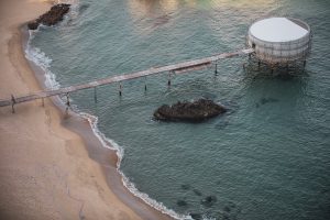 Una imagen aérea de una playa junto al mar, mostrando un muelle que se extiende desde la arena hacia una estructura circular en el agua, con rocas visibles y una posible mancha de derrame de combustible flotando cerca, bajo la luz del atardecer.