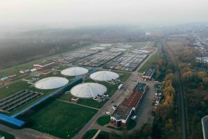 Vista panorámica de una planta de tratamiento con varios reactores anaerobios cubiertos por cúpulas blancas, rodeados de tanques y estructuras industriales, ubicada en un área verde cerca de una vía férrea.